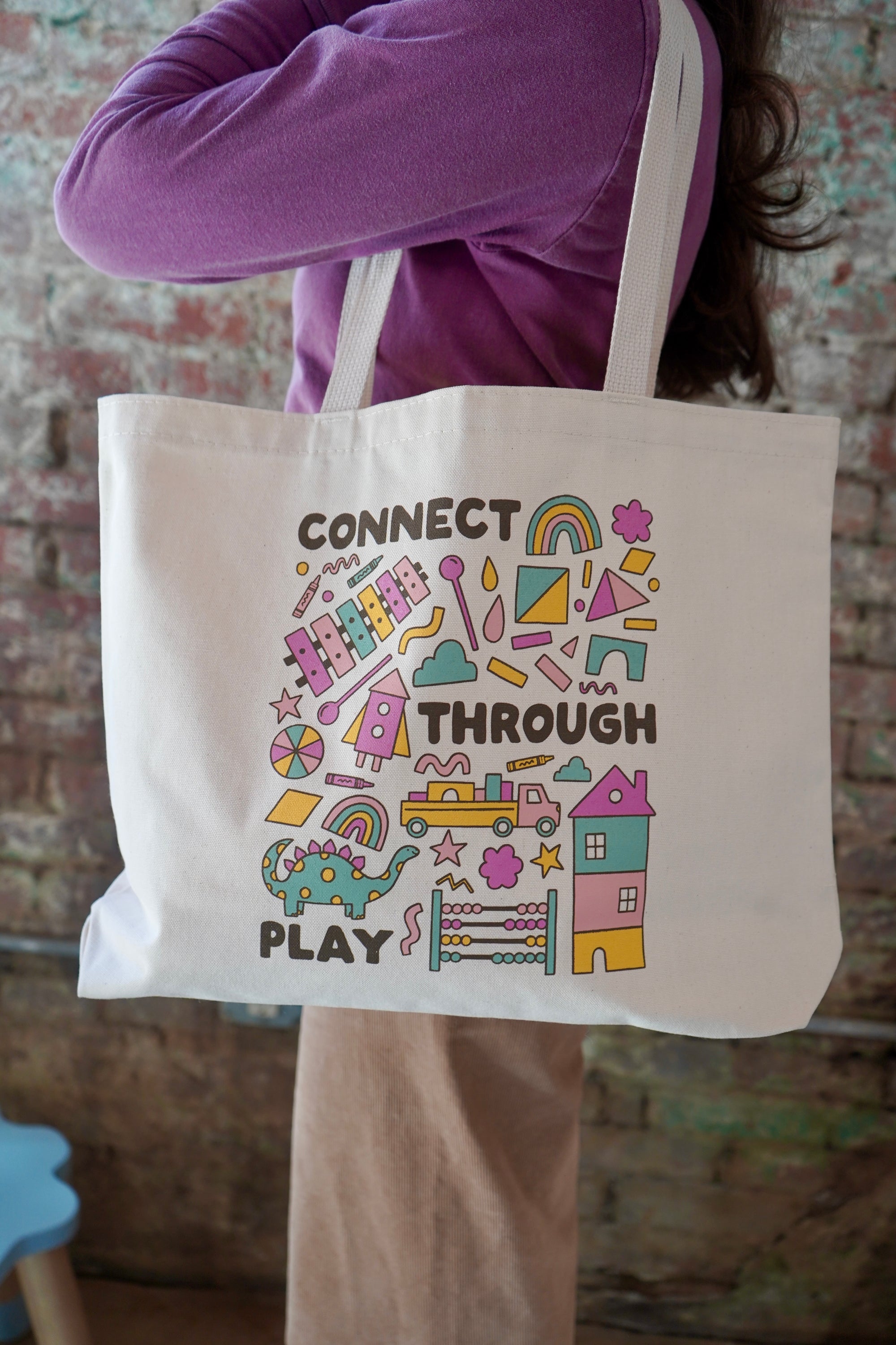 A person wearing a purple sweater is holding a white tote bag featuring colorful graphics and text that says "CONNECT THROUGH PLAY." The background consists of a textured brick wall.