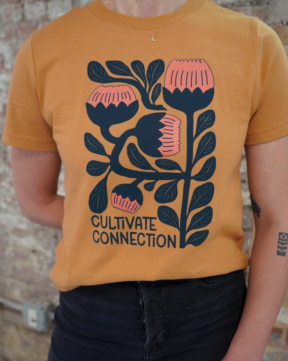 Person wearing a mustard yellow t-shirt with floral design and 'Cultivate Connection' text.