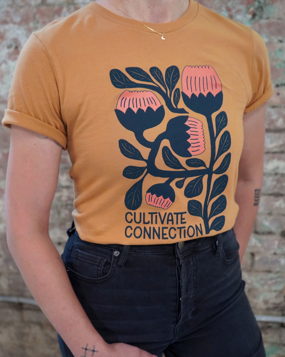 Person wearing a mustard yellow t-shirt with floral design and 'Cultivate Connection' text against a brick wall.