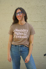 A person wearing a beige t-shirt with 'Disability Rights Are Not Optional' printed in pink text on the front.