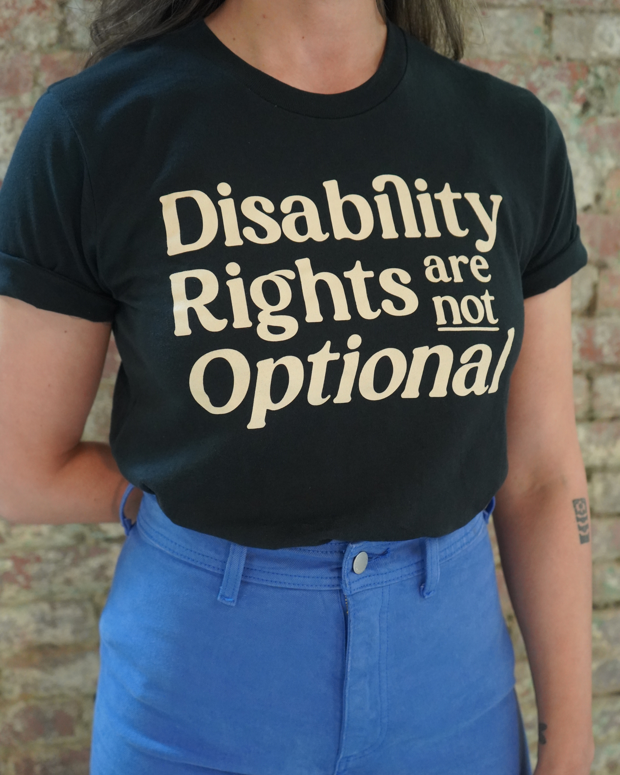 Person wearing a black t-shirt with 'Disability Rights are not Optional' text against a brick wall.