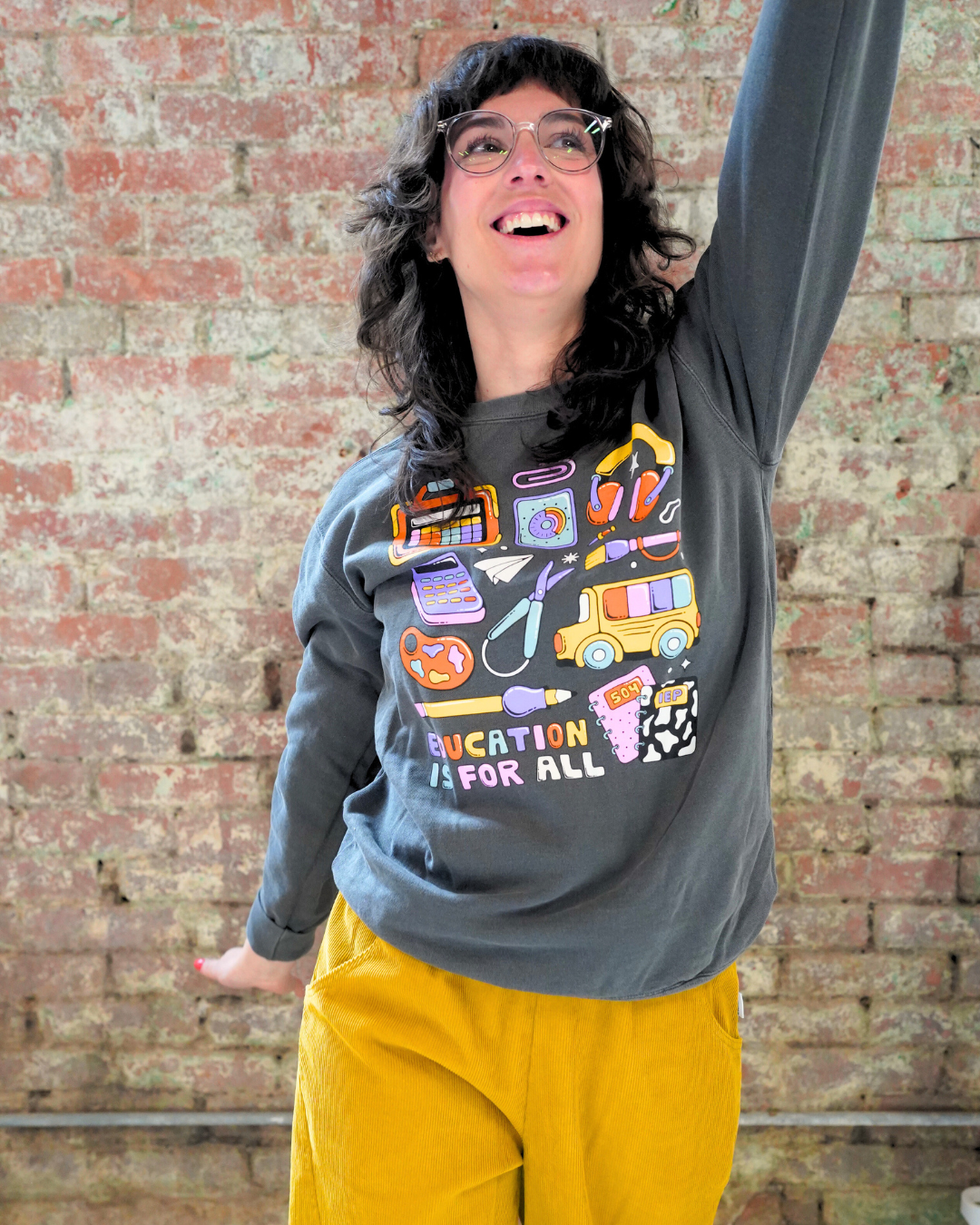 Person wearing a gray sweatshirt with colorful graphics and 'Education is for All' text against a brick wall.