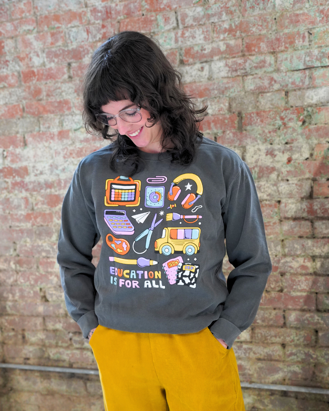 Person wearing a gray sweatshirt with colorful graphics and 'Education is for All' text against a brick wall.