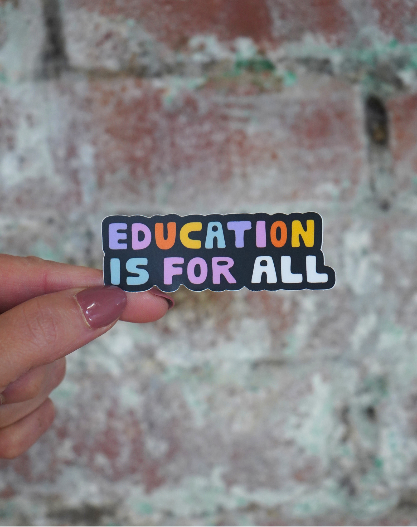 Sticker with 'EDUCATION IS FOR ALL' text held by a hand against a textured wall background