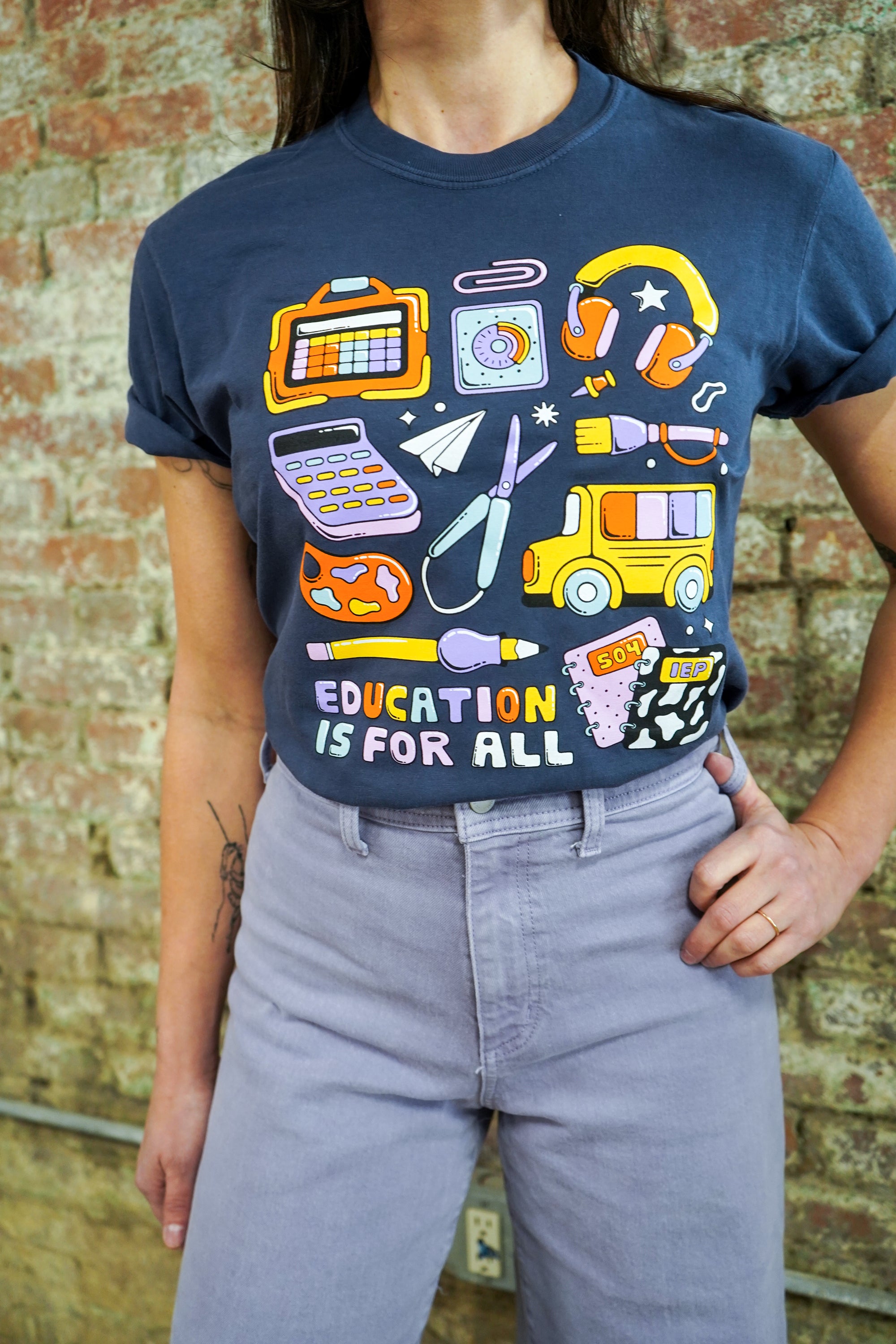 Person wearing a t-shirt with educational graphics and 'Education is for All' text against a brick wall.