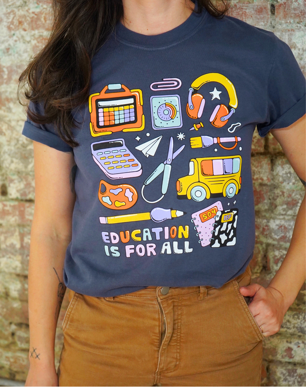 Person wearing a navy blue t-shirt with colorful educational graphics and 'Education is for All' text against a brick wall.