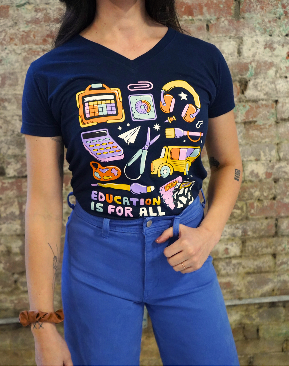 Person wearing a navy blue t-shirt with colorful graphic design and 'Education is for All' text, standing against a brick wall.