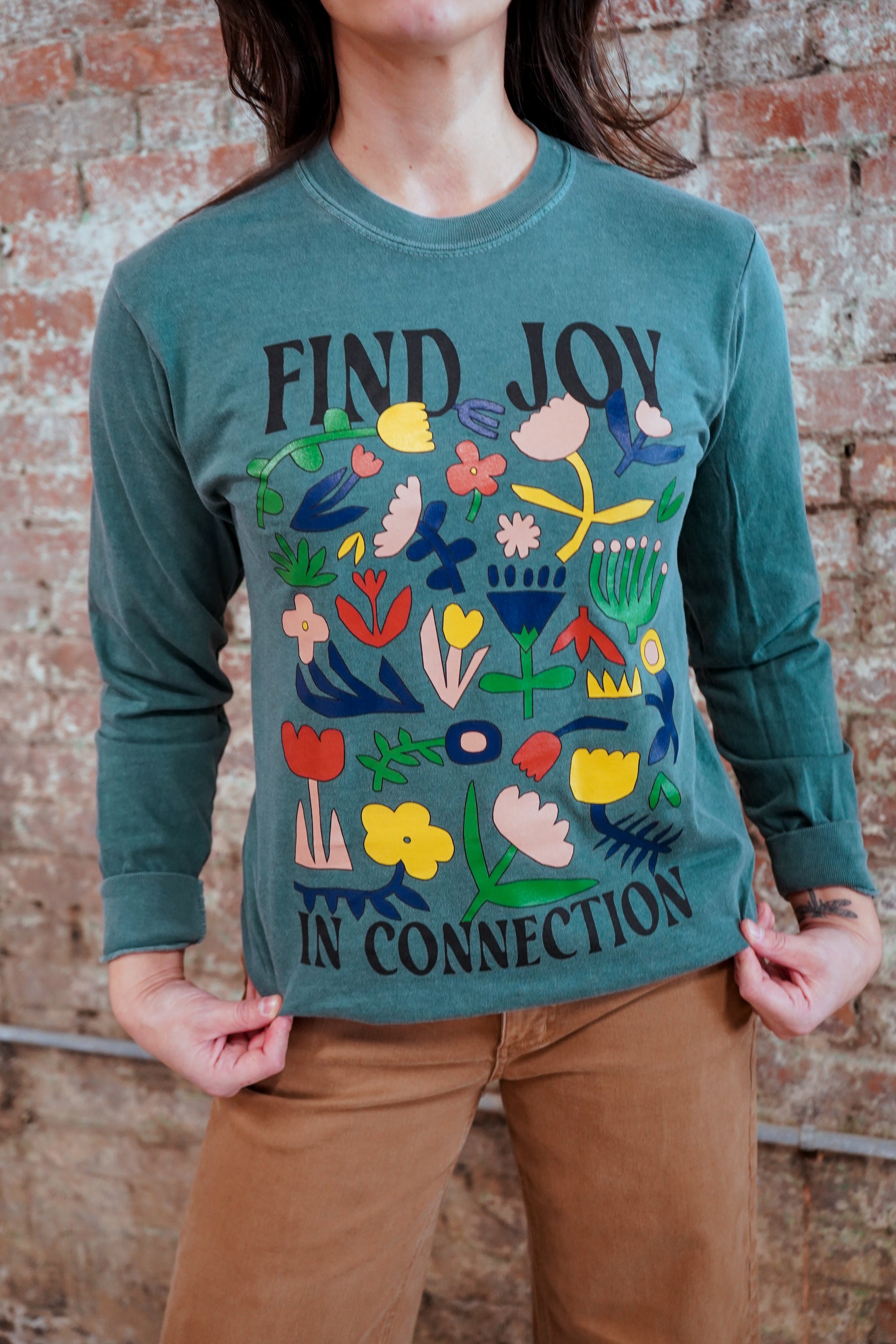 Person wearing a teal long-sleeve shirt with colorful floral design and text against a brick wall.