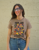 Person wearing a brown t-shirt with colorful design and text against a beige wall.