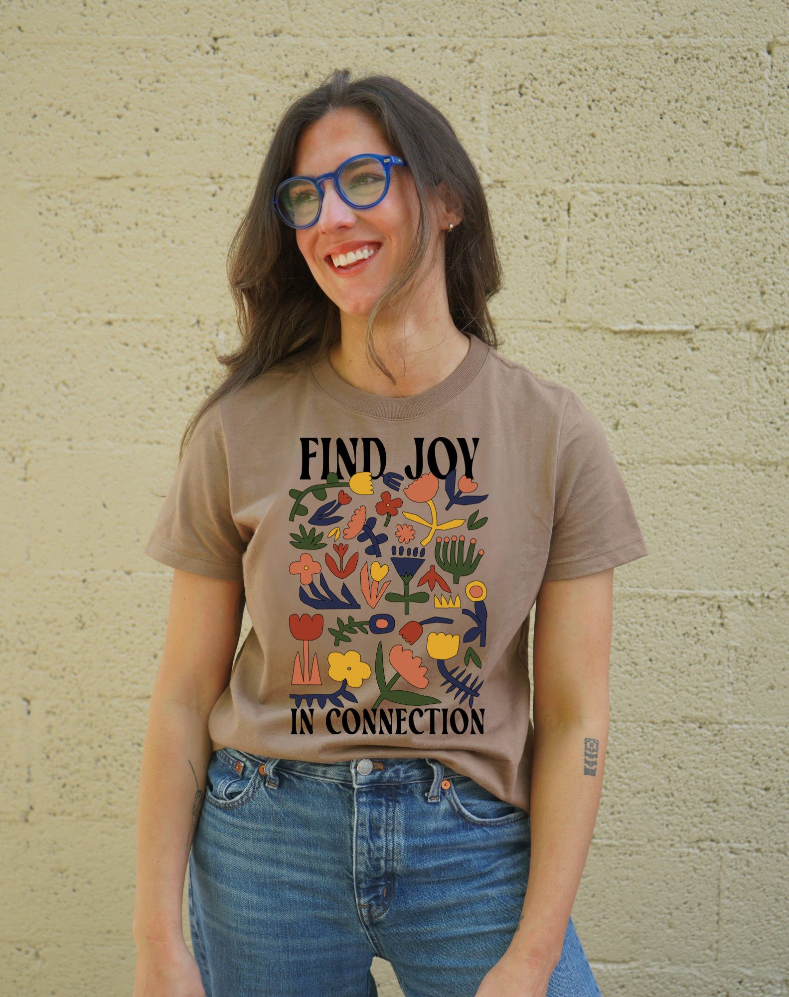 Person wearing a brown t-shirt with colorful design and text against a beige wall.