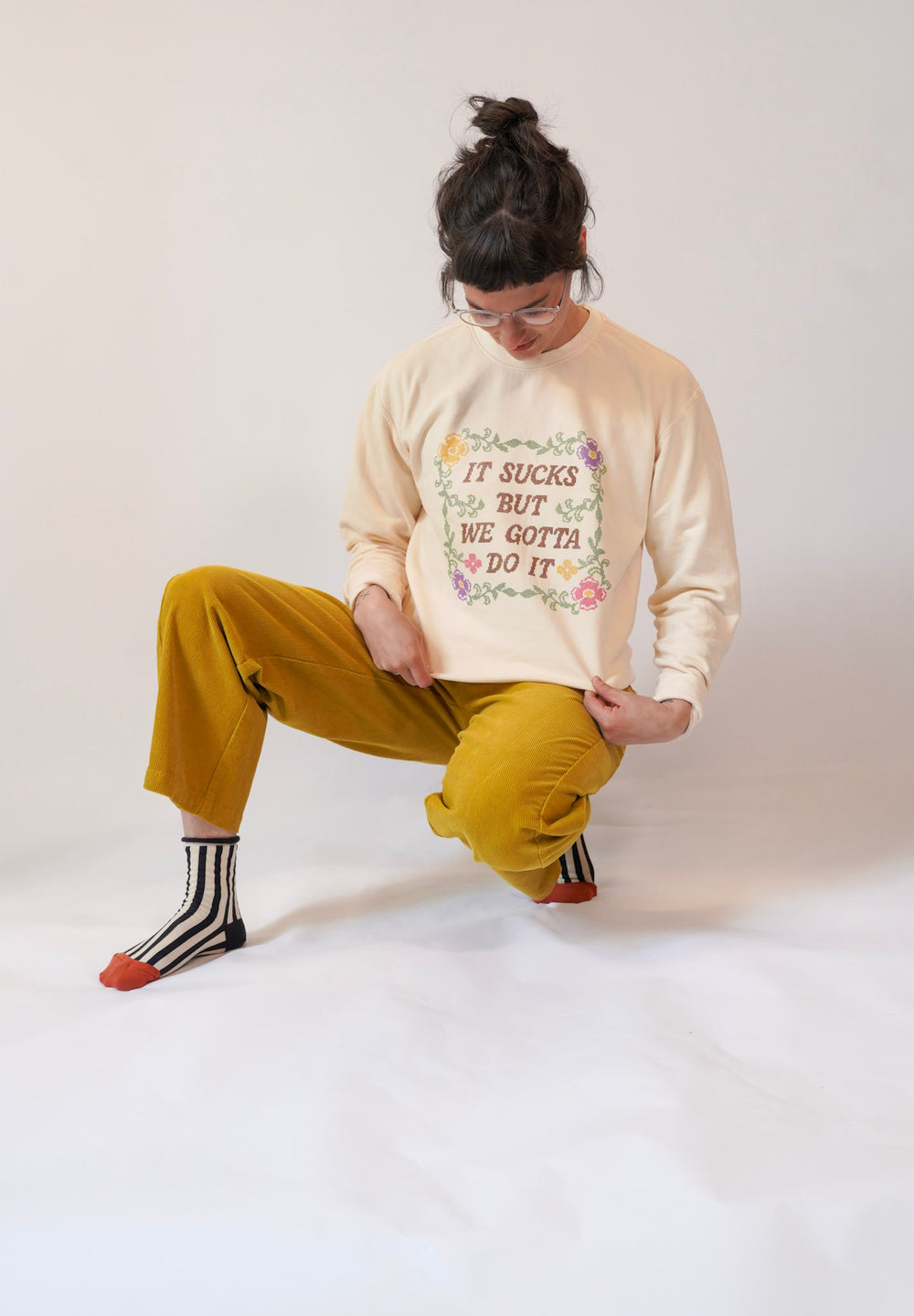 Person wearing a light-colored sweatshirt with text and graphics, mustard yellow pants, and black and white striped socks on a plain background.