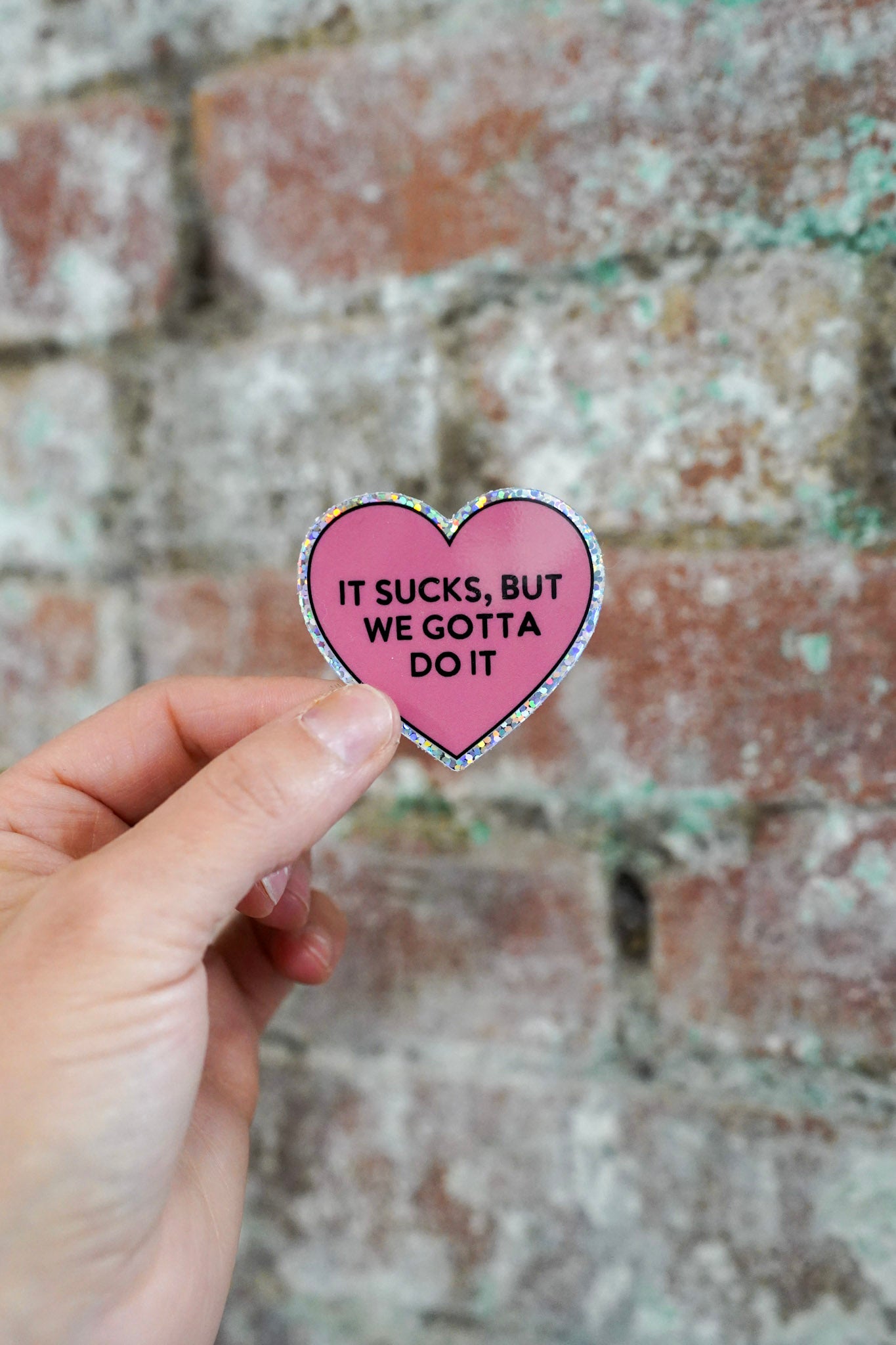 Heart-shaped sticker with text held in front of a brick wall