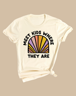 A beige colored t-shirt with a graphic that reads 'MEET KIDS WHERE THEY ARE' with a colorful semi-circle behind the text.