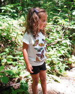 Play Spark Notice The Beauty In Our Differences Children's Tee in white, featuring colorful butterfly graphics on the front. The tee is shown worn by a child in a natural setting, complemented by black shorts.