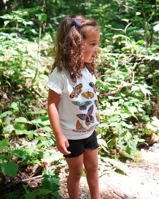Play Spark Notice The Beauty In Our Differences Children's Tee in white, featuring colorful butterfly graphics on the front. The tee is shown worn by a child in a natural setting, complemented by black shorts.