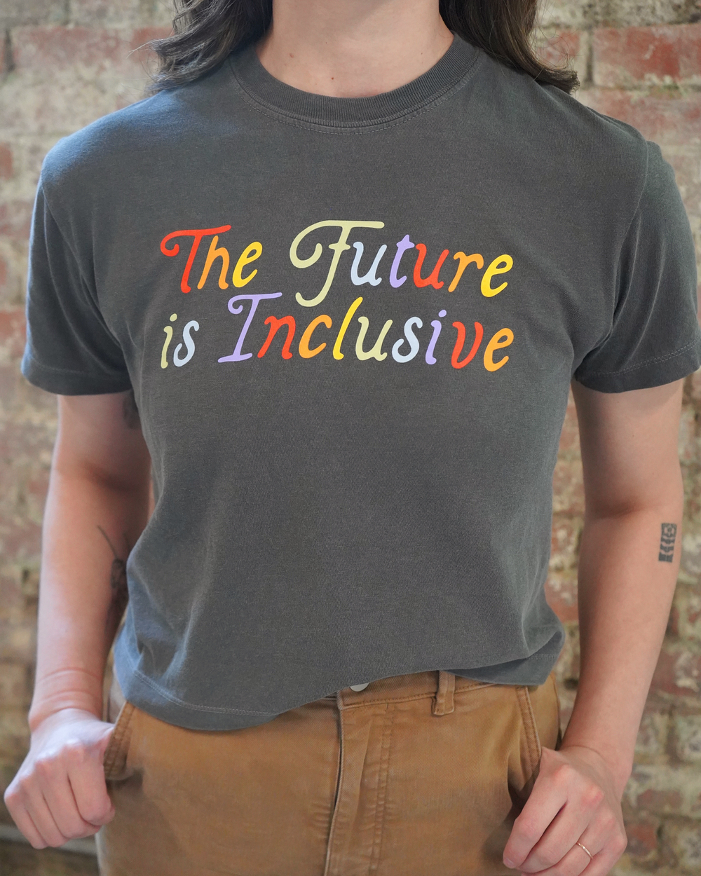 Person wearing a gray t-shirt with 'The Future is Inclusive' in colorful letters against a brick wall.