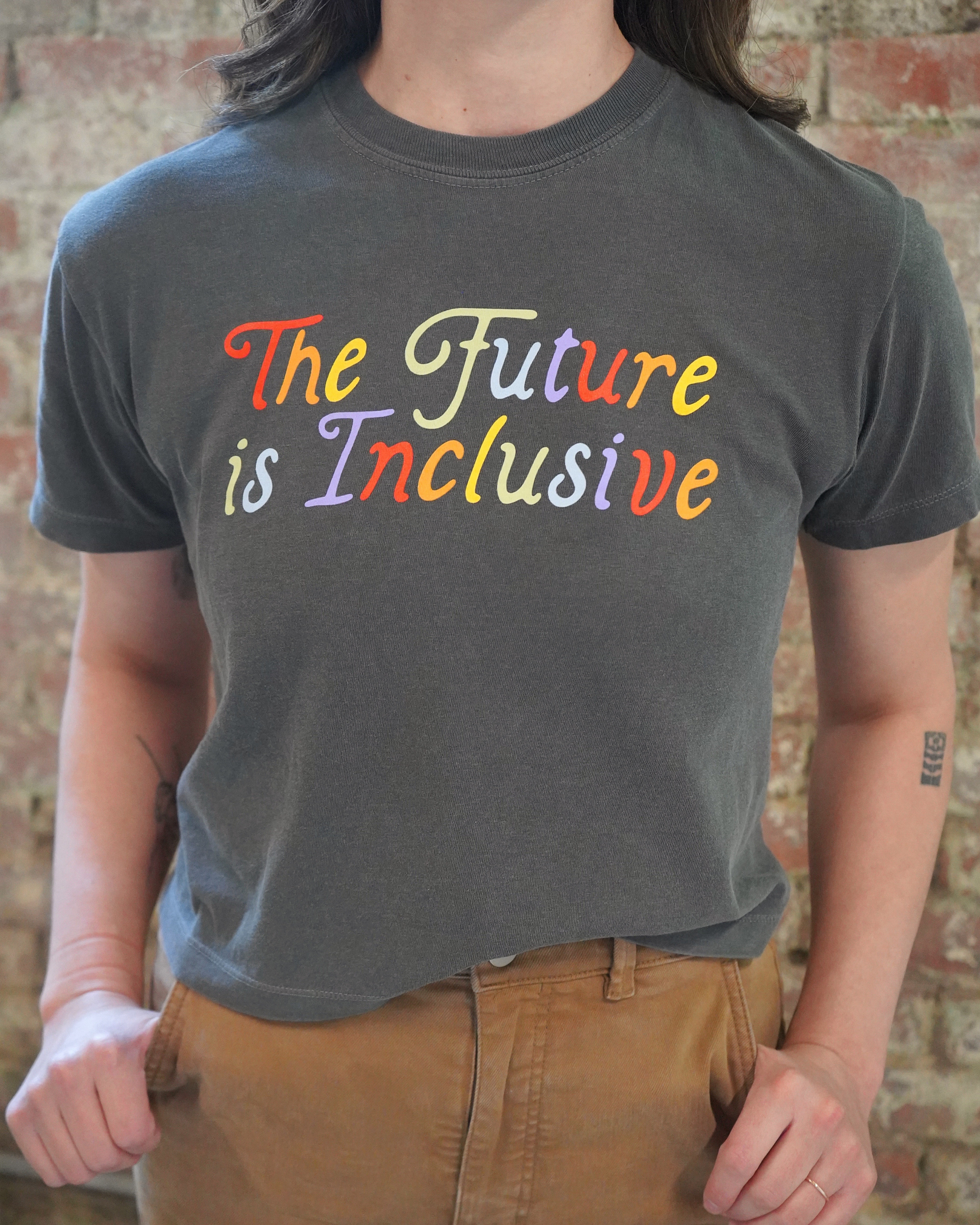 Person wearing a gray t-shirt with 'The Future is Inclusive' in colorful letters against a brick wall.