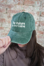 Person wearing a green cap with 'The Future is Inclusive' text against a brick wall.
