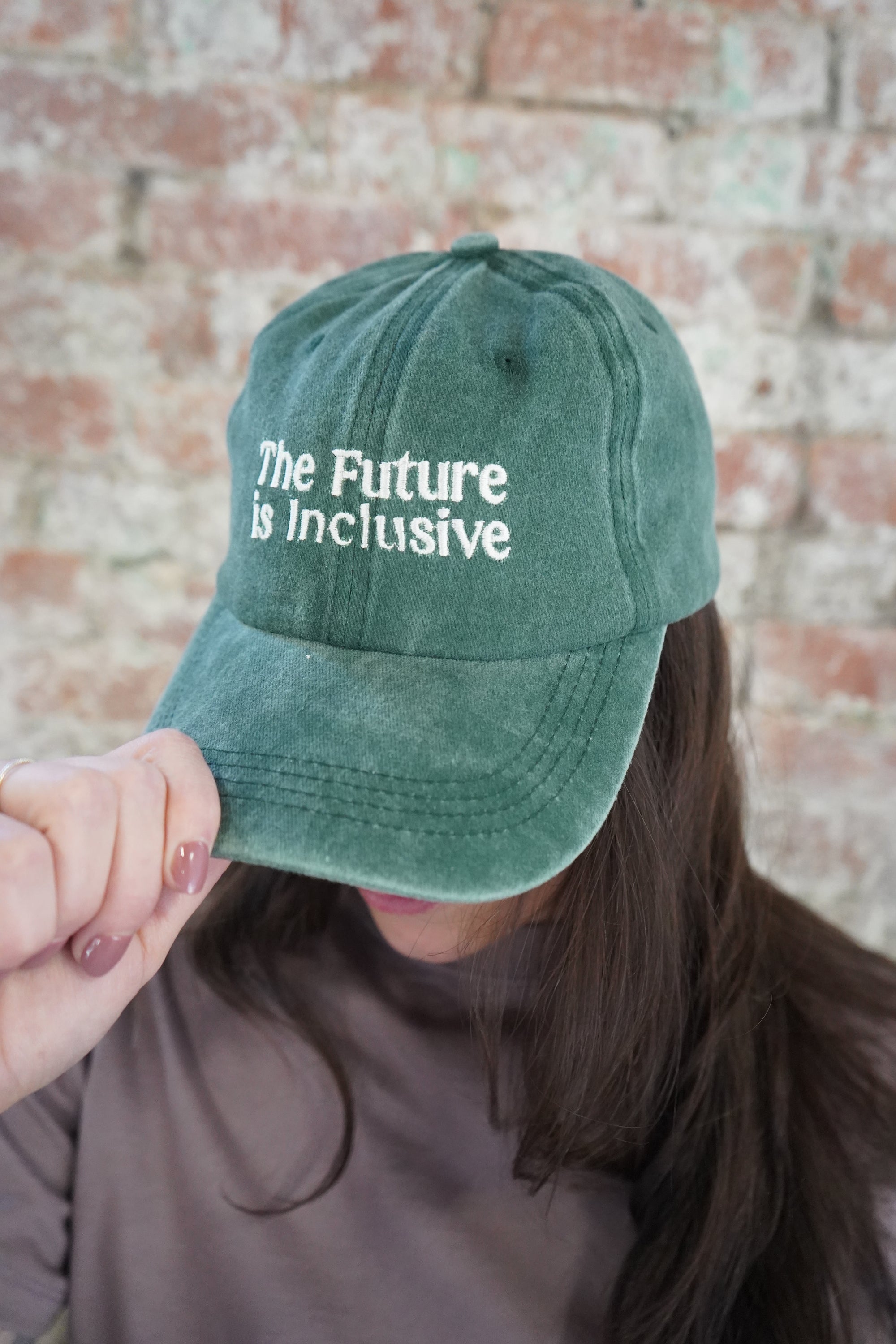 Person wearing a green cap with 'The Future is Inclusive' text against a brick wall.