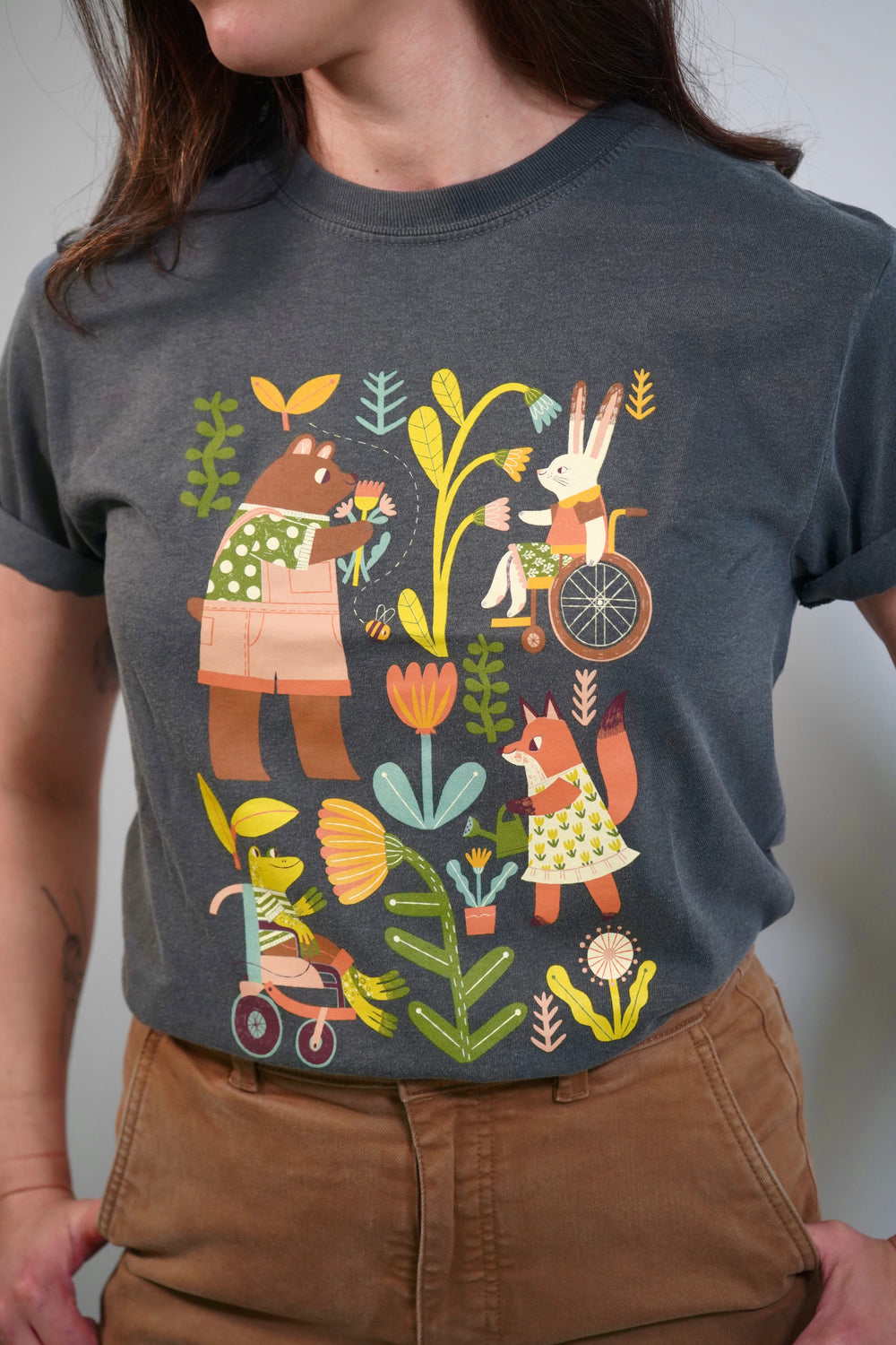 A person wearing a gray t-shirt featuring a colorful illustration of various anthropomorphic animals. A bear, a rabbit in a wheelchair, and a fox are surrounded by plants and flowers, showcasing a friendly, inclusive scene.