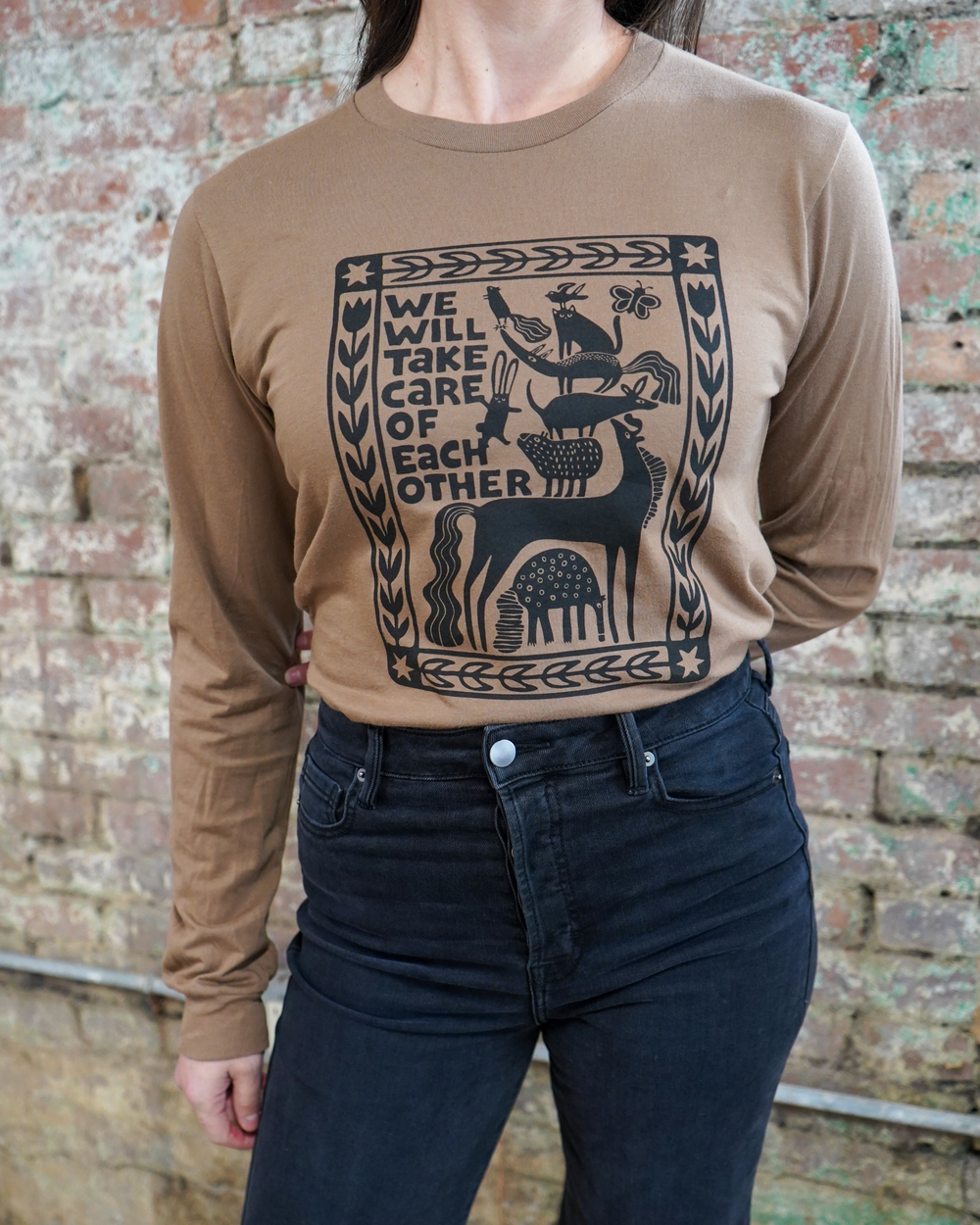 Person wearing a brown long-sleeve shirt with a graphic design and text against a brick wall.
