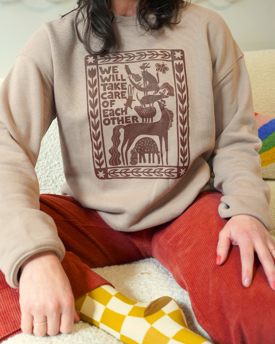 Person wearing a beige sweatshirt with a graphic design and text, sitting on a colorful rug.