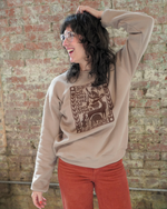 Person wearing a beige sweatshirt with a graphic design against a brick wall.