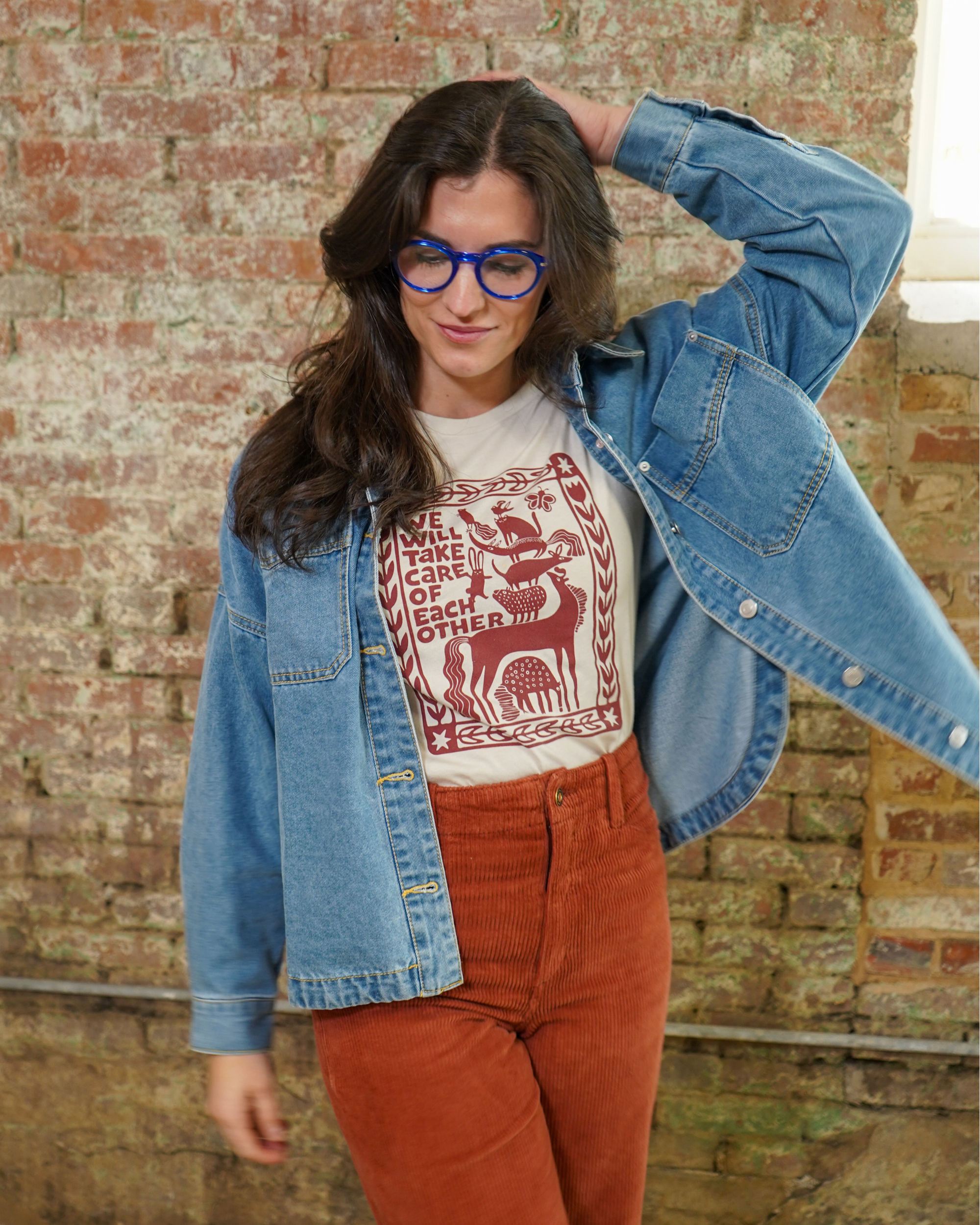 Woman wearing a denim jacket, white shirt with red design saying "We Will Take Care of Each Other", and red pants against a brick wall.