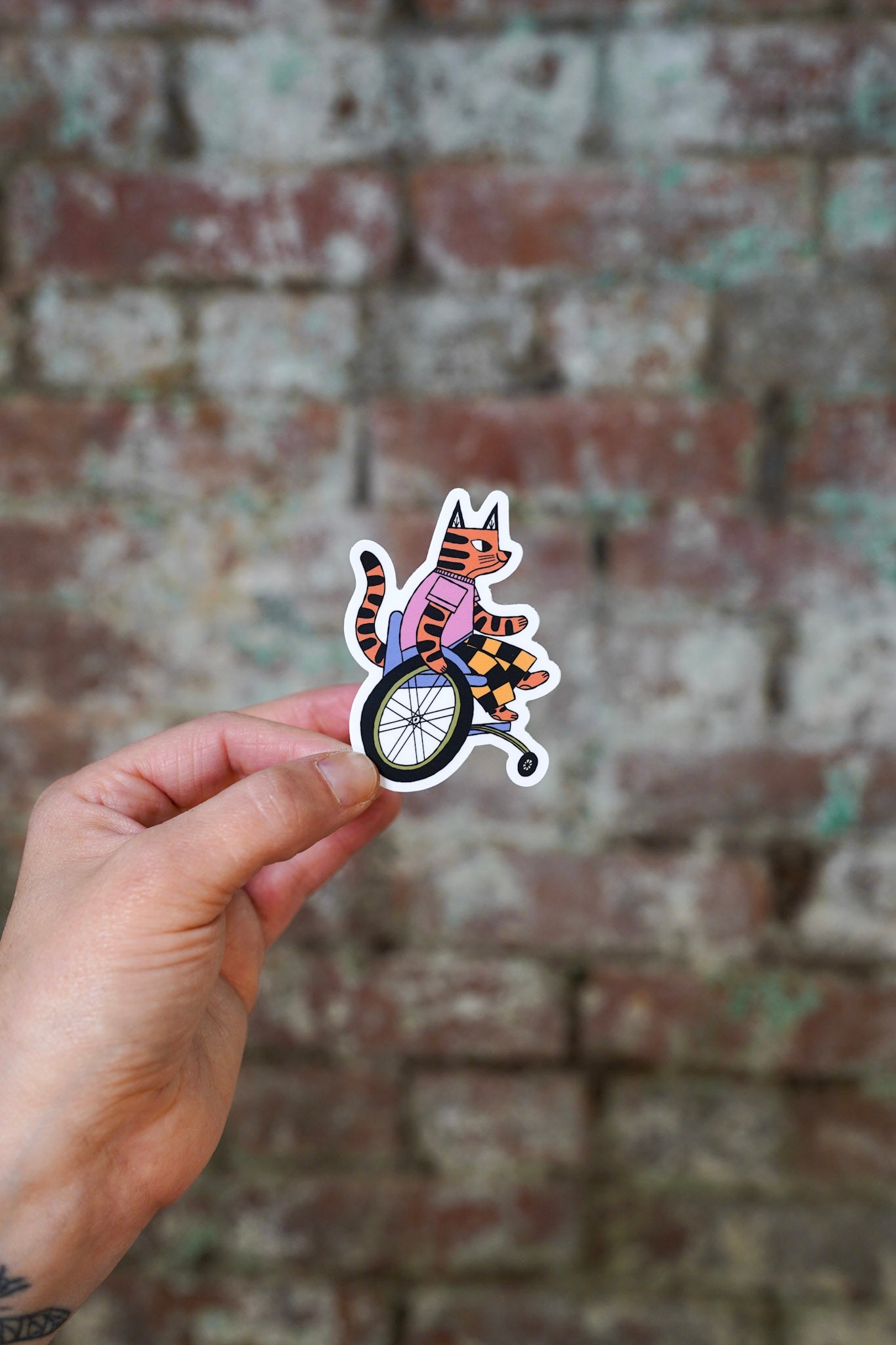 Hand holding a sticker of a tiger in a wheelchair against a brick wall.